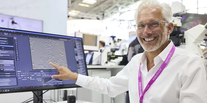 A smiling man wearing glasses and a white shirt points to a microscope image on a computer screen at Analytica USA, surrounded by lab equipment and other attendees.