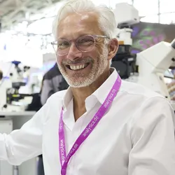 A smiling man wearing glasses and a white shirt points to a microscope image on a computer screen at Analytica USA, surrounded by lab equipment and other attendees.