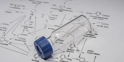 A laboratory sample vial resting on a sheet of chemical structures and formulas, representing analytical chemistry and mass spectrometry research.