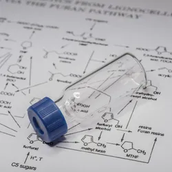 A laboratory sample vial resting on a sheet of chemical structures and formulas, representing analytical chemistry and mass spectrometry research.