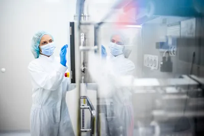 A scientist in a cleanroom suit, face mask, and gloves adjusts stainless steel bioprocessing equipment, illustrating process intensification in a sterile biomanufacturing environment.