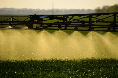 Close-up of a crop sprayer boom applying a fine mist of agricultural treatments to a green field at sunset, representing the real-world residues analyzed by the QECh*All mega-method for food safety monitoring.