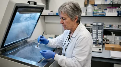 A histology specialist guides a thin tissue section onto a slide at an open cryostat, with slides and a sublimation apparatus on the bench alongside.